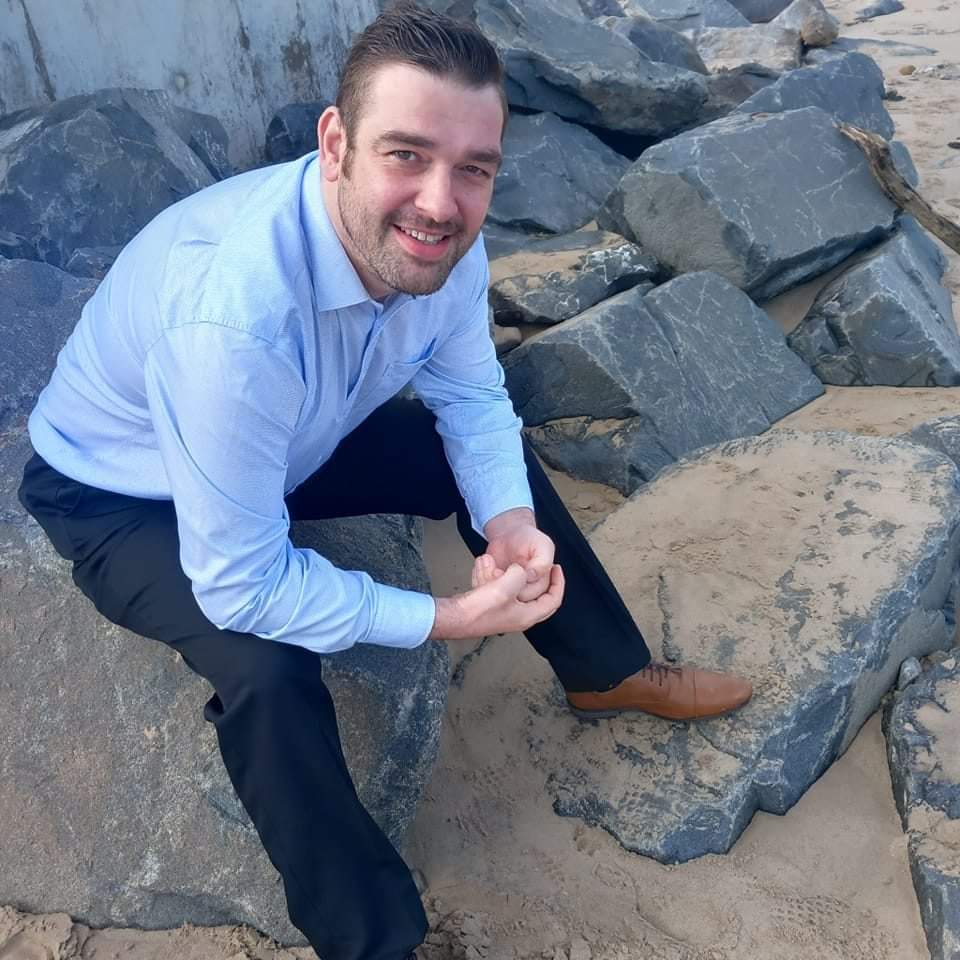 John Nicholson ADI Approved driving Instructor wearing a suit with a blue dress shirt, brown shoes, and jeans smiling at the camera