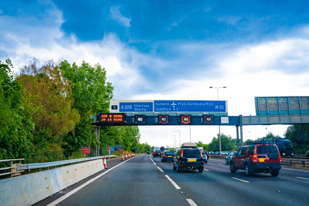 A UK motorway with digital speed limit signs, route directions, and vehicles driving on the left lane.